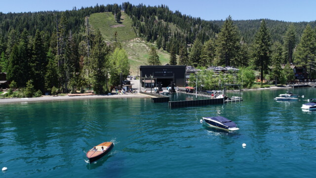 Homewood Marina, tucked beneath its namesake ski slope. Aerial view of the Homewood Marina building at the shoreline, with two boats cruising the turquoise water and a forested, ski-slope-lined mountain rising behind.