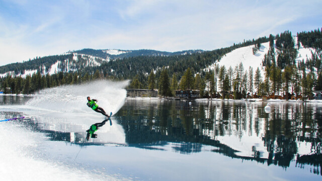 A striking mirror shot of the same winter water skiing run. A wetsuit-clad water skier in a neon vest carves a hard turn on mirror-calm water, throwing a large spray, with snowy mountains and lakeside cabins reflected behind him.