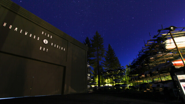 Homewood Marina under a starry night sky. A star-filled night sky stretches above the "Homewood High & Dry Marina" boathouse and illuminated boat storage racks, silhouetted pine trees standing between them.