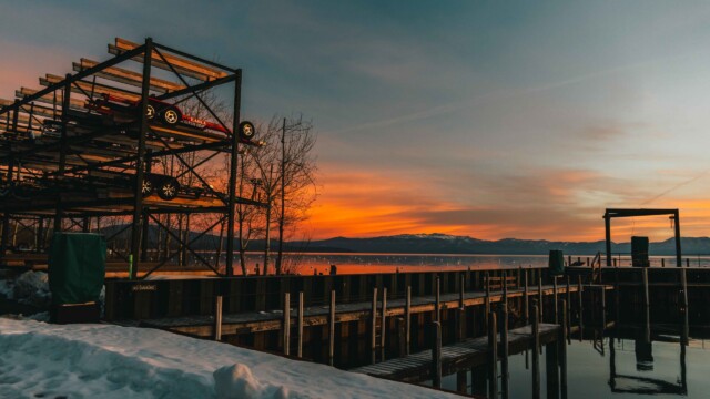 Winter gold — a Tahoe sunset from the Homewood dock. A fiery winter sunset over Lake Tahoe viewed from Homewood Marina's snow-covered dock, with empty boat racks silhouetted against the orange sky.