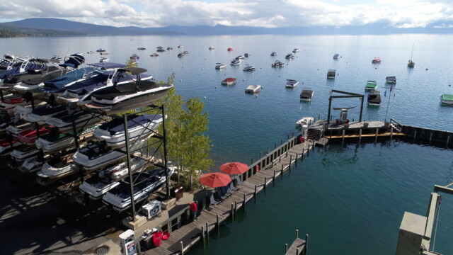 A busy marina on Lake Tahoe, boats stacked at the dock and dozens more moored offshore. Aerial view of a lakeside marina with stacked boat storage, a long wooden dock with red umbrellas, and dozens of boats moored on the open lake.